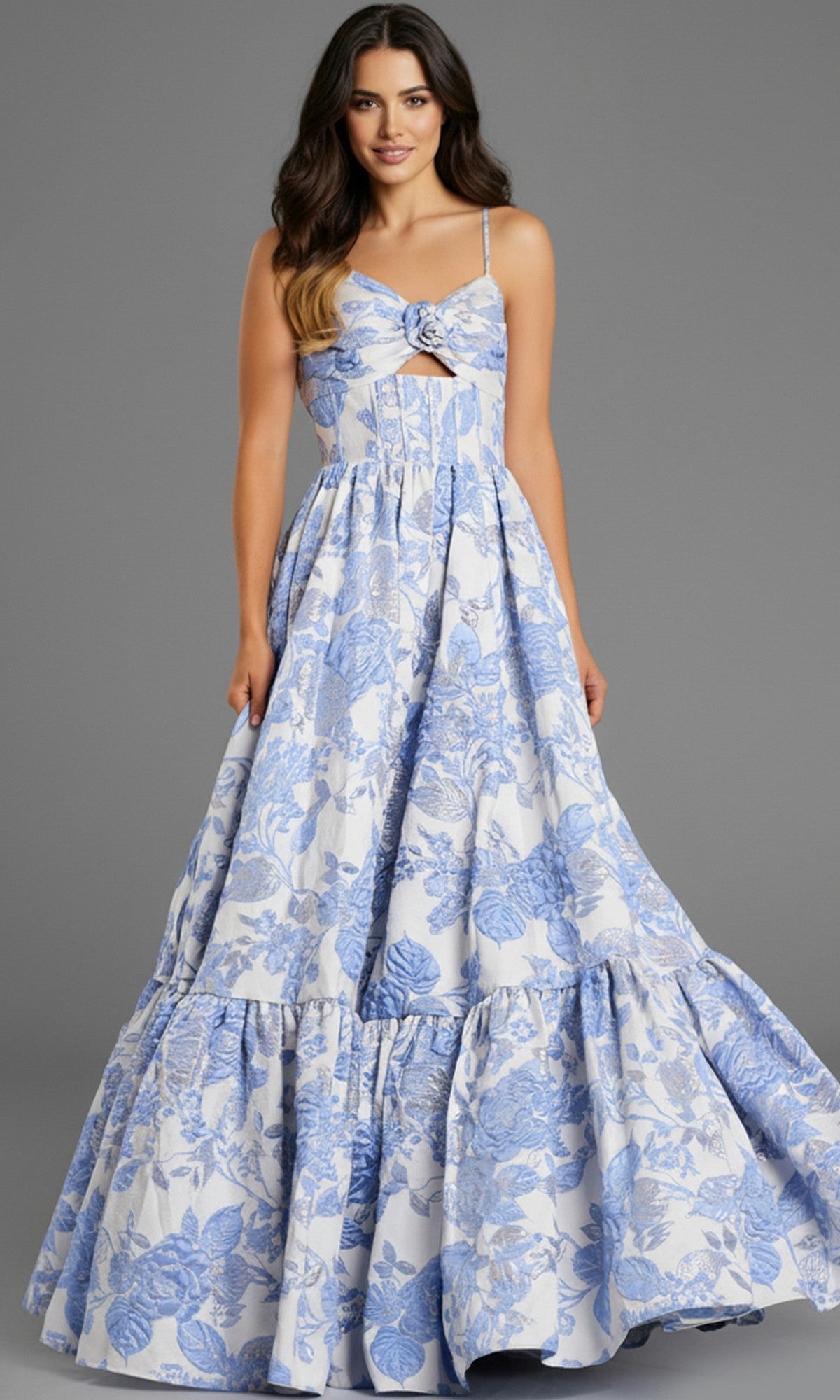 A woman stands against a plain gray background wearing the Jovani 47328 long prom dress, featuring a sleeveless floral jacquard design with blue patterns and an elegant A-line skirt.